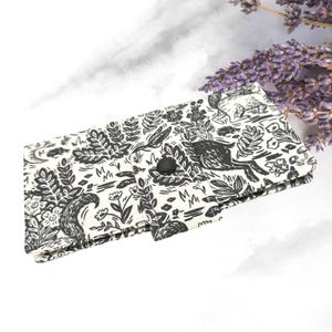 May include: A white and black patterned wallet featuring a forest scene with a fox, flowers, and foliage. The wallet has a black button closure. A sprig of purple lavender is in the background.