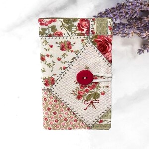 May include: A rectangular fabric cover featuring a patchwork design of cream, green, and red floral patterns. The cover is secured with a bright red circular button and a white elastic loop, showcasing a detailed rose print and small floral motifs.