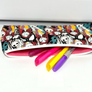 May include: A rectangular pencil case with a zipper, featuring a colorful pattern of characters. The interior is lined with pink fabric, and several markers in pink, yellow, and purple are partially visible.