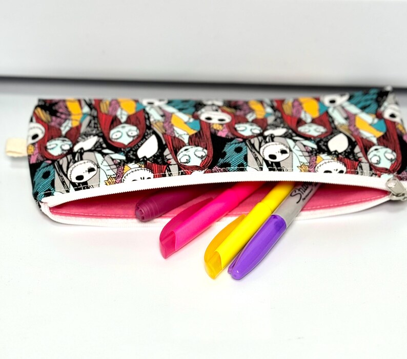 May include: A rectangular pencil case with a zipper, featuring a colorful pattern of characters. The interior is lined with pink fabric, and several markers in pink, yellow, and purple are partially visible.