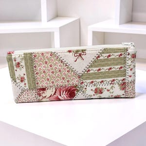 May include: A rectangular pencil case with a patchwork design. The case features a variety of floral patterns in shades of pink, green, and cream. A small red bow is stitched near the zipper. The case is approximately 8 inches long.