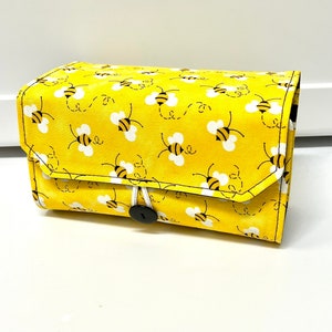 Peut inclure: Une pochette en tissu jaune avec une fermeture à bouton blanc. La pochette est décorée d'un motif répétitif d'abeilles noires et jaunes.