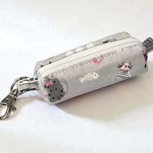 May include: A small, rectangular gray fabric pouch with a white zipper and a silver-colored metal clasp. The pouch is decorated with a repeating pattern of cartoon cats, fish bones, and hearts. The pouch is approximately 5 inches long.