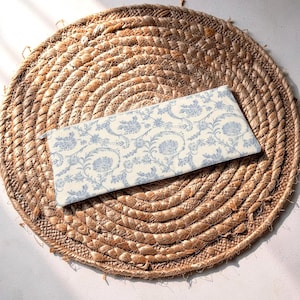May include: A rectangular pencil case with a blue floral pattern on a cream background rests on a round, woven, brown placemat. The pencil case has a zipper closure and is made of fabric. The placemat is made of natural fibers.