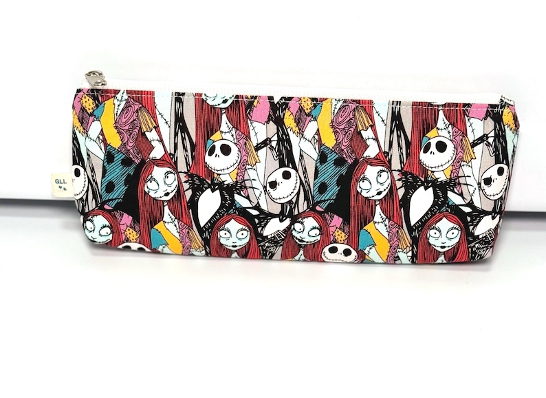 May include: A rectangular pencil case with a white zipper and a colorful pattern featuring characters from "The Nightmare Before Christmas." The design includes Sally, Jack Skellington, and other characters in a repeating pattern.