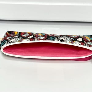 May include: A zippered pencil case with a black, white, and teal pattern featuring a character design. The interior is a bright pink, and the zipper is white with a silver pull. The case is open, revealing the interior.