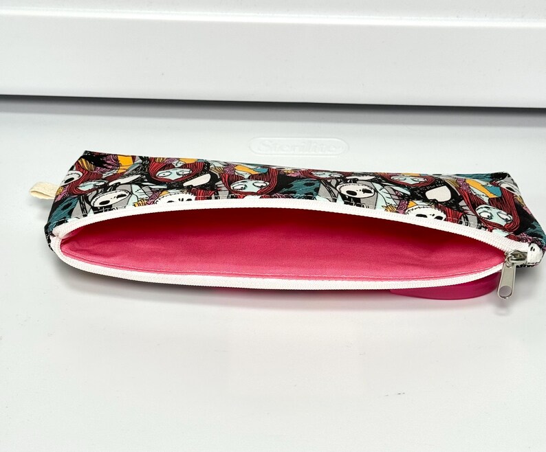 May include: A zippered pencil case with a black, white, and teal pattern featuring a character design. The interior is a bright pink, and the zipper is white with a silver pull. The case is open, revealing the interior.