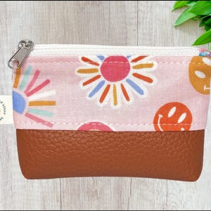 May include: A small, rectangular coin purse with a brown leather bottom and a pink fabric top. The fabric features a repeating pattern of colorful flowers and smiley faces. A silver zipper and a small tag that says "Grandmas Little Lily Handmade" are visible.
