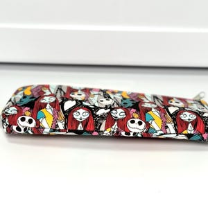 May include: A rectangular pencil case with a zipper closure. The case is covered in a colorful pattern featuring characters from The Nightmare Before Christmas, including Sally and Jack Skellington. The background is black, with accents of red, yellow, and blue.