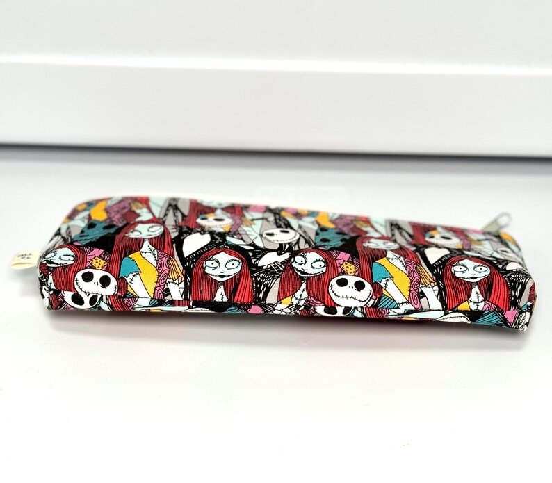May include: A rectangular pencil case with a zipper closure. The case is covered in a colorful pattern featuring characters from The Nightmare Before Christmas, including Sally and Jack Skellington. The background is black, with accents of red, yellow, and blue.