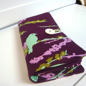 May include: A purple fabric wallet with a floral pattern featuring green birds and a white button closure.