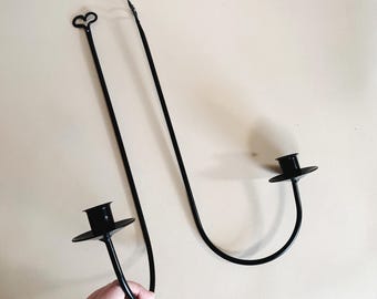 May include: Black metal candle holder with two arms, each with a round candle cup. The holder is designed to be mounted on a wall.