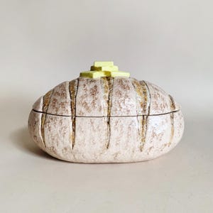 May include: A ceramic bread-shaped lidded container. The off-white container has vertical brown stripes and a textured surface. A small, yellow, geometric shape sits on top of the lid. The container is on a white background.
