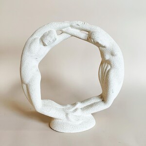May include: White ceramic sculpture of two figures forming a circle. The figures are stylized and abstract, with smooth, rounded forms.