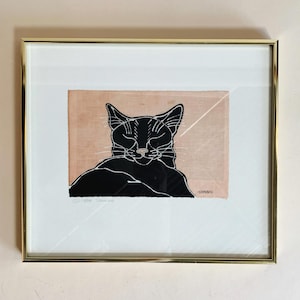 May include: A framed black and white print of a sleeping cat. The cat is outlined in black and the background is a light pink. The print is signed "TAMAKO" in the bottom right corner.