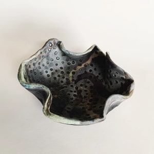 May include: A ceramic bowl with a unique, irregular shape and a speckled, dark gray glaze. The bowl has a pattern of small holes throughout the surface.