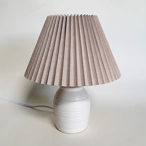 May include: A white ceramic table lamp with a pleated beige linen lampshade. The lamp base has a textured, ribbed design.