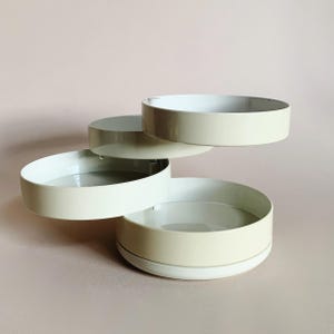 May include: A cream-colored, tiered serving tray with four circular levels. The trays are arranged in a staggered, spiral design, with a light beige exterior and a white interior. The background is a solid, pale pink.