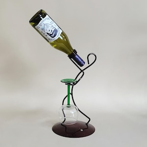 May include: A whimsical black metal wine bottle holder in the shape of a person, holding a wine bottle and a wine glass. The holder has a green accent and a dark brown base. The wine bottle is green and has a label.