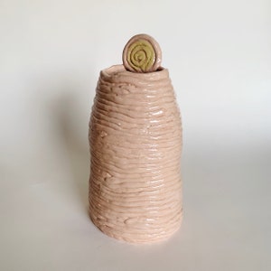 May include: A pink ceramic vase with a textured, ribbed surface. The vase has a small, green and yellow ceramic swirl on top.