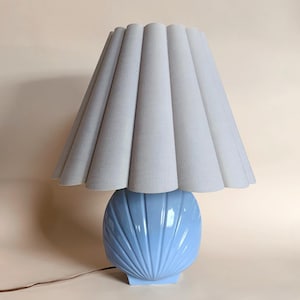May include: A light blue ceramic table lamp with a seashell-shaped base and a pleated, light gray lampshade. The lamp has a cord and plug. The lamp is a decorative lighting fixture.