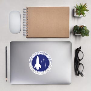 Puede incluir: Una pegatina para portátil con el logotipo de la British Interplanetary Society. El diseño incluye un cohete blanco, tres estrellas y el texto "British Interplanetary Society" y "From Imagination to Reality" sobre un fondo azul.