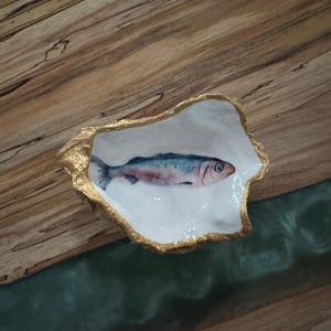 May include: A decorative, white oyster shell-shaped dish with a gold-painted rim. The dish features a watercolor painting of a fish with blue, pink, and silver scales. The dish is set on a wooden surface.