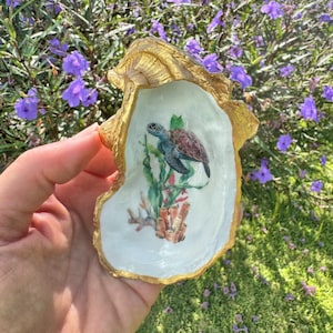 May include: A gold-painted oyster shell with a painted image of a sea turtle swimming in coral. The shell is a decorative dish.