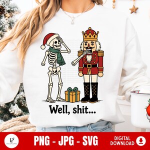 Może przedstawiać: Biała bluza z długim rękawem z grafiką szkieletu w czapce Mikołaja i zielonym szaliku, stojącego obok dziadka do orzechów. Szkielet trzyma rękę na czole. Poniżej znajduje się pudełko z prezentem oraz napis "Well, shit..."