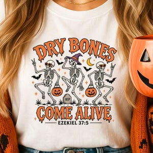 May include: A white t-shirt featuring a graphic of three dancing skeletons with pumpkins, bats, and a crescent moon. The text reads "DRY BONES COME ALIVE" in orange, with "EZEKIEL 37:5" printed below in black. The skeletons wear sunglasses and hats.