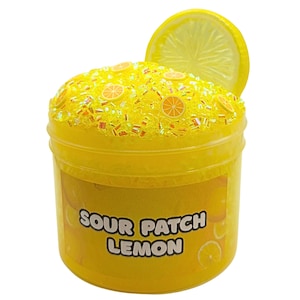 May include: A yellow plastic container filled with yellow slime, decorated with small orange slices and glitter. A large lemon slice is placed on top. The text "Sour Patch Lemon" is printed on the container.