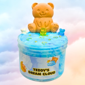 Peut inclure: Slime bleu et blanc avec un ours en peluche brun assis sur le dessus. Le slime a des paillettes dorées et des étoiles. Le pot a une étiquette qui dit "Teddy's Dream Cloud".