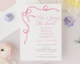 Op de afbeelding: Een roze en witte bruidsdouche-uitnodiging met een aquarelillustratie van een roze lint dat in een strik is gebonden. De tekst luidt "She's tying the knot" en bevat de naam van de bruid, datum, tijd en locatie. De uitnodiging bevat ook de tekst "Registered at Crate & Barrel and Anthropologie."