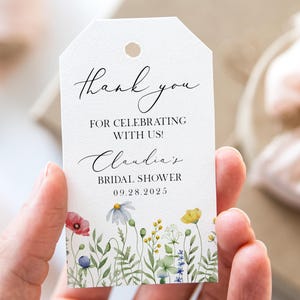 May include: A white tag with a floral border and the words "thank you" in elegant script. The tag reads "For celebrating with us! Claudia's Bridal Shower 09.28.2025". The tag is held in a hand, with a gift box and ribbon in the background.