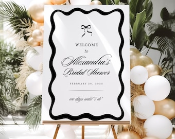 May include: A rectangular white welcome sign with a thick black wavy border, featuring a black bow icon and text reading: WELCOME to Alexandra's Bridal Shower, FEBRUARY 24, 2025, and "one days until 'i do'". The sign rests on a wooden easel.
