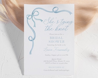 Op de afbeelding: Een lichtblauwe bruidsdouche-uitnodiging met een aquarel lintontwerp. De uitnodiging luidt "She's tying the knot" en bevat details over het evenement, waaronder de naam van de bruid, datum, tijd, locatie en RSVP-informatie. De uitnodiging bevat ook de tekst "Registered at Crate & Barrel and Anthropologie."