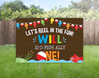 May include: A brown wooden yard sign with the text "LET'S REEL IN THE FUN! WILL IS O-FISH-ALLY ONE!" features colorful fish and bobber decorations. The sign is placed in a grassy yard with a fence and trees in the background.