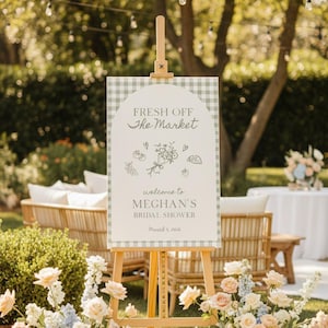 May include: An outdoor scene featuring a wooden easel holding a sign with the text "FRESH OFF the MARKET" and "MEGHAN'S BRIDAL SHOWER". The sign has a gingham border and floral illustrations. Flowers and seating are visible.