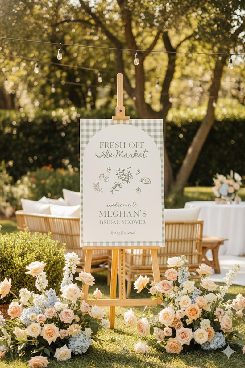 May include: An outdoor scene featuring a wooden easel holding a sign with the text "FRESH OFF the MARKET" and "MEGHAN'S BRIDAL SHOWER". The sign has a gingham border and floral illustrations. Flowers and seating are visible.