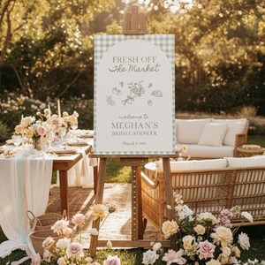 May include: A bridal shower setup outdoors, featuring a sign on an easel with the text "Fresh Off The Market" and "Welcome to Meghan's Bridal Shower". The scene includes floral arrangements, a wicker sofa, and soft lighting, creating a warm atmosphere.