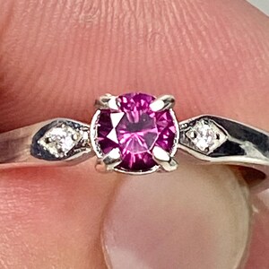 Op de afbeelding: Een zilveren ring met een ronde, donkerroze edelsteen omgeven door kleinere, heldere edelstenen.