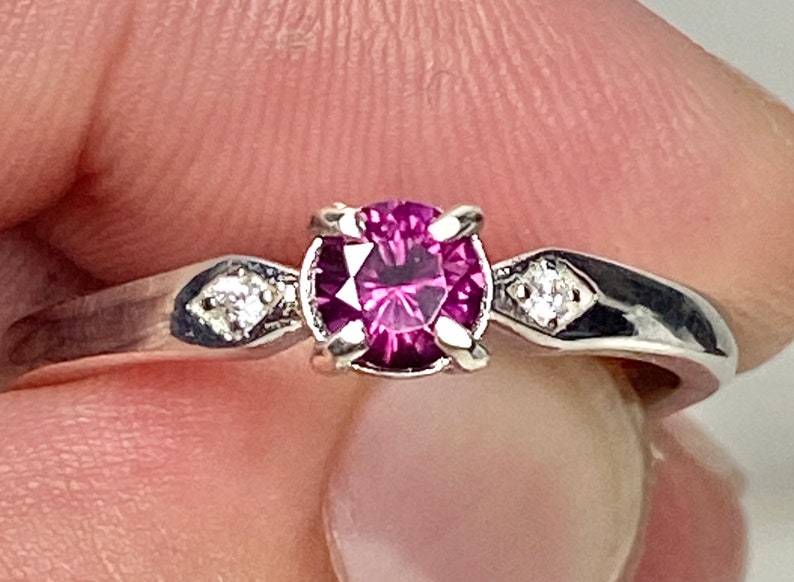 Op de afbeelding: Een zilveren ring met een ronde, donkerroze edelsteen omgeven door kleinere, heldere edelstenen.