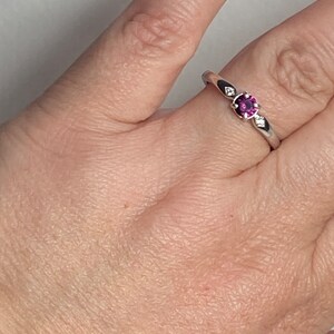Op de afbeelding: Een zilveren ring met een kleine, ronde, paarse edelsteen in het midden. Twee kleinere, heldere edelstenen zijn aan weerszijden van de middelste steen gezet.