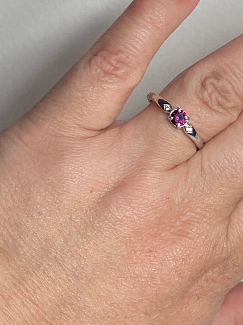 Op de afbeelding: Een zilveren ring met een kleine, ronde, paarse edelsteen in het midden. Twee kleinere, heldere edelstenen zijn aan weerszijden van de middelste steen gezet.