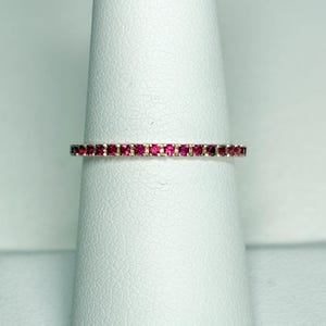 May include: A thin, delicate eternity band ring featuring a continuous row of small, round, vibrant pinkish-red gemstones set in a polished metallic band. The ring is designed with a slim profile, showcasing a uniform arrangement of stones.