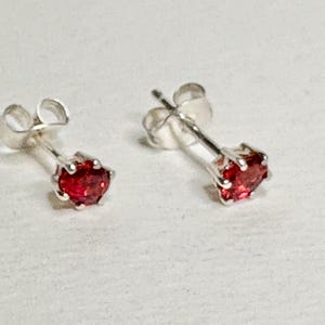 May include: A pair of stud earrings with bright red gemstones. The earrings have a silver-colored metal setting and posts. The gemstones are round and faceted, catching the light. The earrings are displayed on a light-colored surface.
