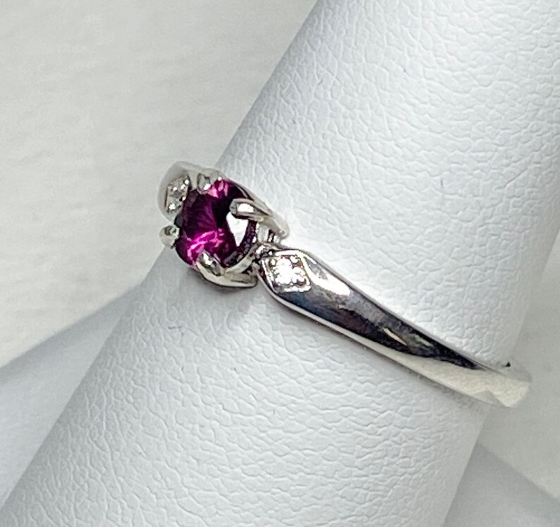 Op de afbeelding: Een zilveren ring met een enkele, ronde, donkerpaarse edelsteen omringd door kleinere, heldere edelstenen.