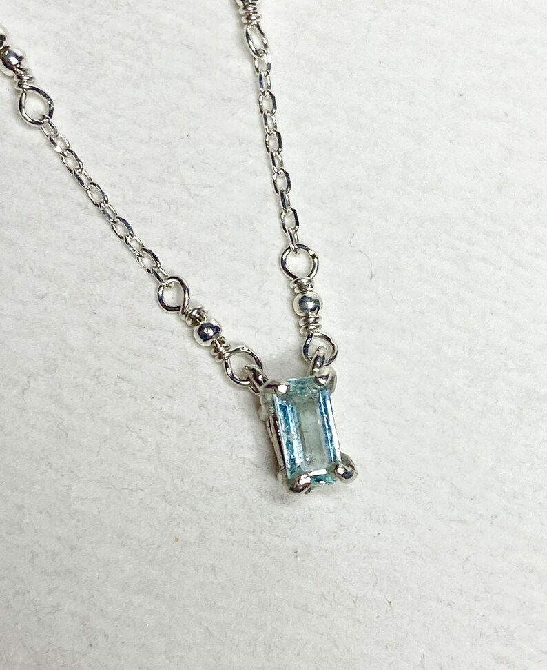 May include: A delicate silver chain necklace with a small, rectangular aquamarine gemstone pendant.