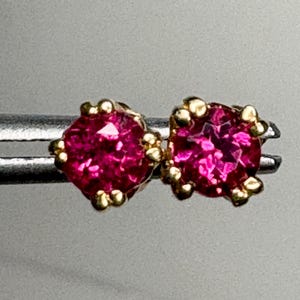May include: A pair of round, faceted, vibrant pink gemstones set in four-prong gold metal settings. The two circular stones are held side-by-side by a silver-toned metal tool, showcasing the bright magenta color and the reflective, cut surfaces.