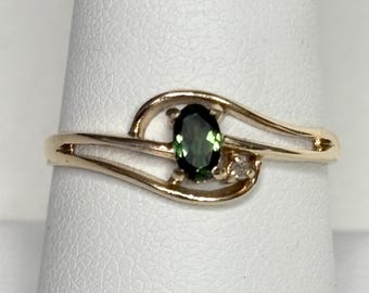 May include: A gold ring featuring an oval-cut, dark green gemstone as the primary focal point. The ring has a delicate, swirling design with a small, round diamond accent. The band is a simple, polished gold.